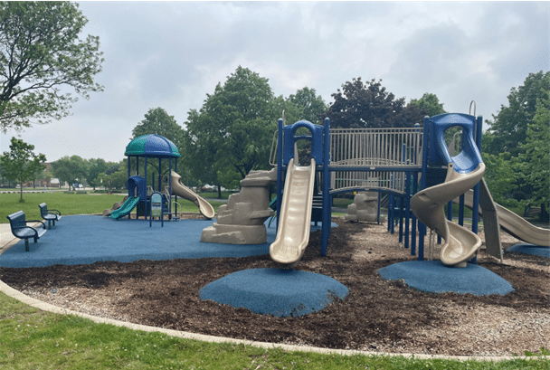A large blue and tan commercial playground structure with multiple slides, climbing walls, and integrated safety surfacing in a public park.
